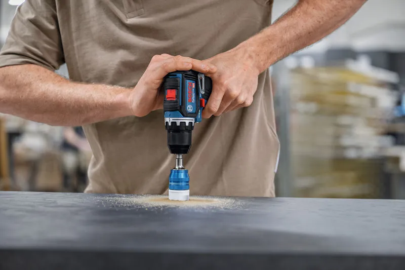 Person, der betjener en akku-boremaskine med et hulsavtilbehør på en mørk overflade.